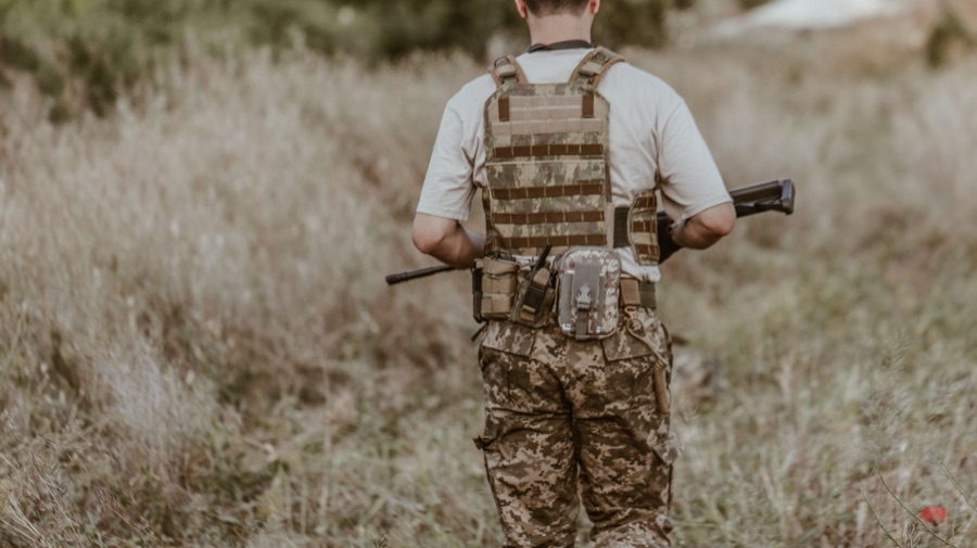 Airsoft player from behind wearing a coyote tan plate carrier and battle belt with magazine pouches in a grass field