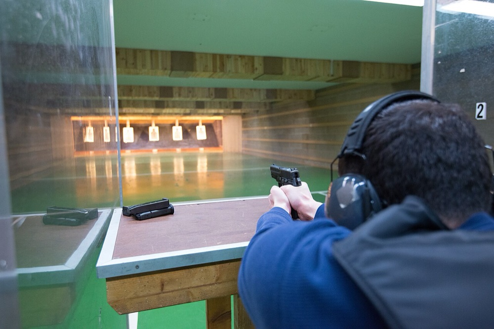 Shooter aiming a pistol at an indoor range with spare magazines on the bench