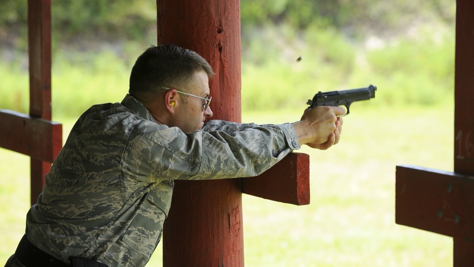 A U.S. Air Force officer wearing yellow foam earplugs fires a Beretta M9 from behind a wooden post on an outdoor range, with a spent case flying above the pistol