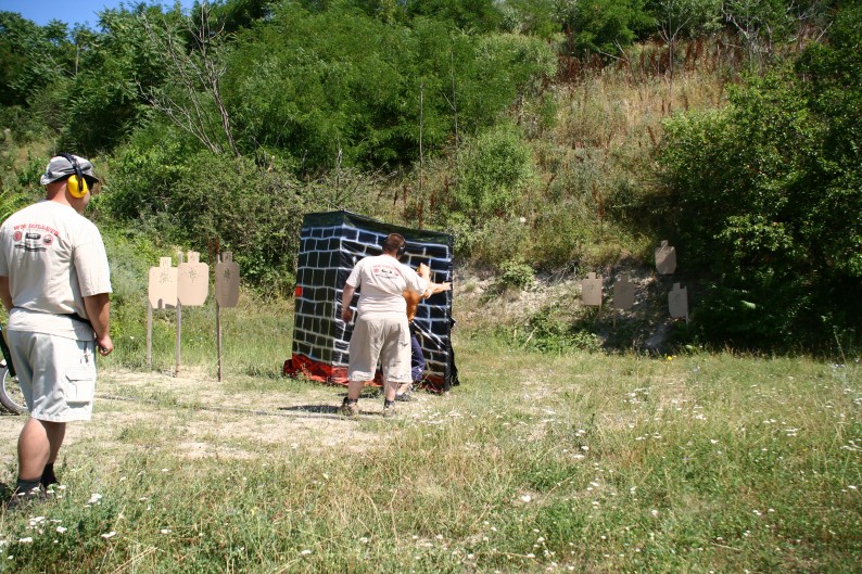 A club-level IDPA stage — cardboard threats spread across several distances, a fabric barricade, and the next shooter watching from behind the line