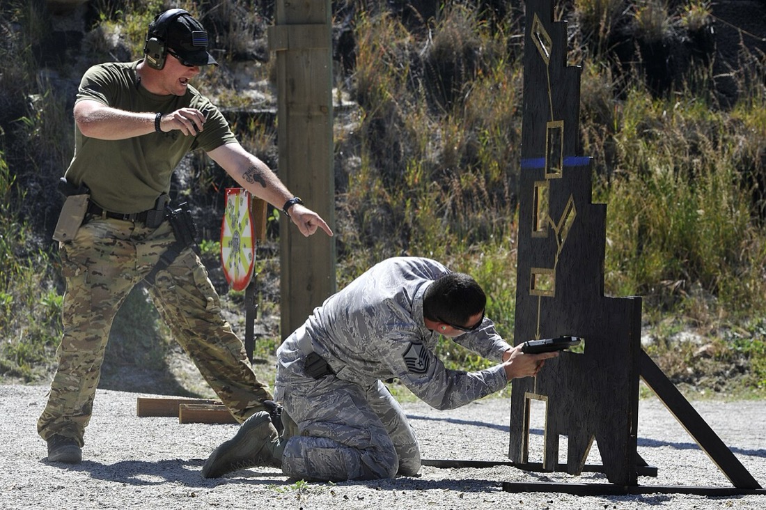 A Safety Officer standing beside a kneeling shooter and calling the stage — the SO’s commands run the whole sequence from “Load and make ready” to “Range is clear”