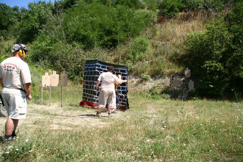 IDPA 比賽進行中——選手繞著掩體打關卡，遠處立著瓦楞紙人形靶