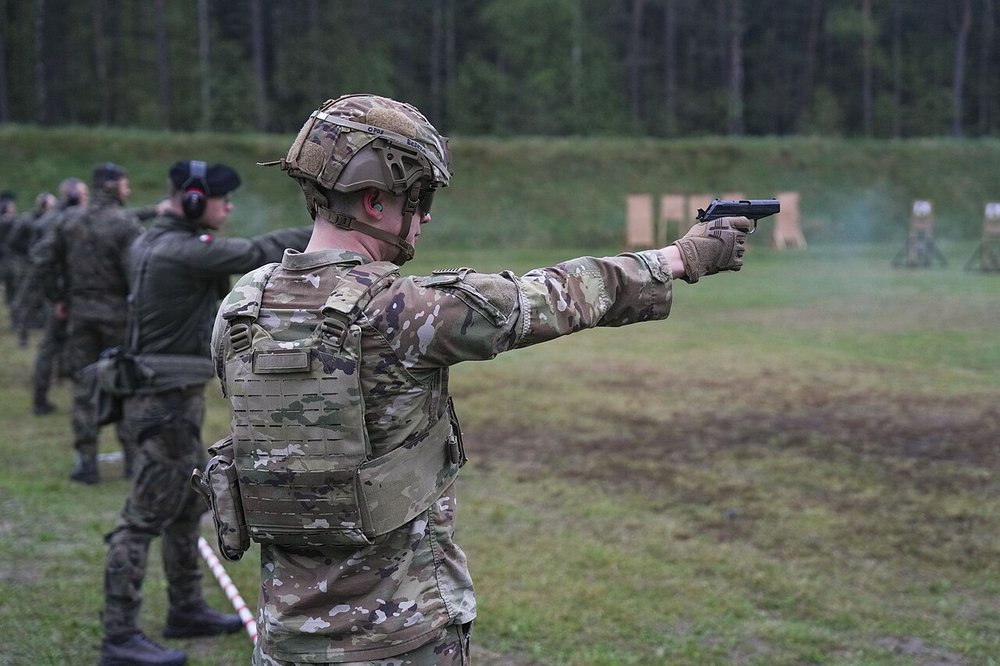 U.S. Army soldier firing a P-83 pistol one-handed at a Polish marksmanship competition, with a line of shooters also firing one-handed behind him