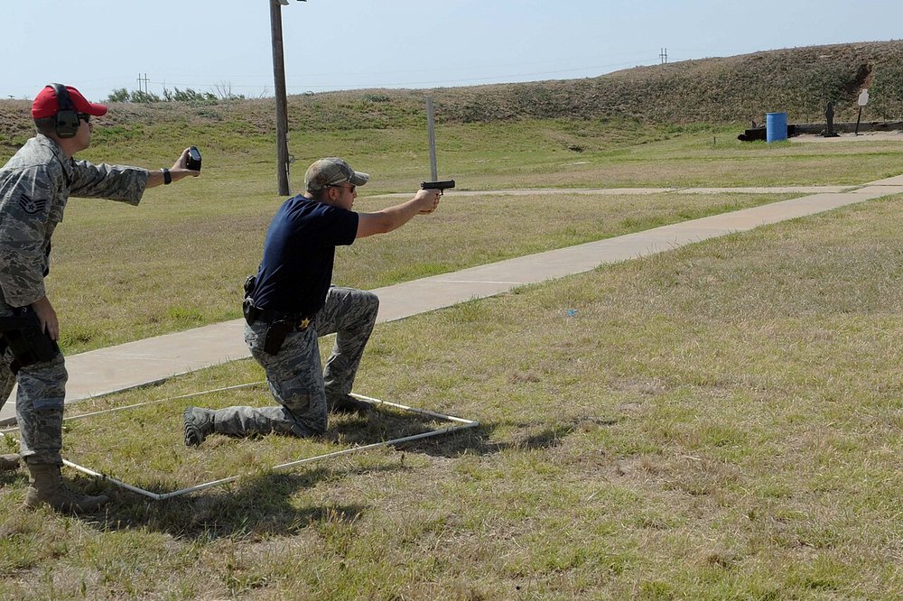 Range officer holding a handheld shot timer behind a kneeling shooter firing an M9 pistol during a timed police week shootout