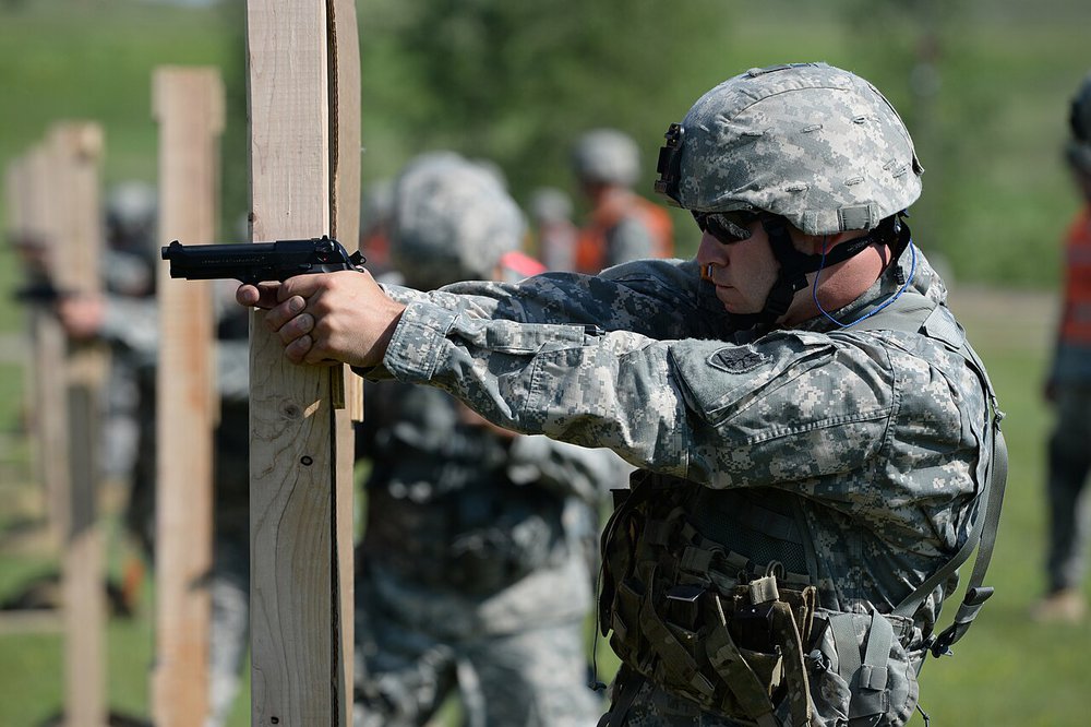 National Guard soldier beside a wooden barricade preparing to shoot an M9 pistol with his non-dominant hand