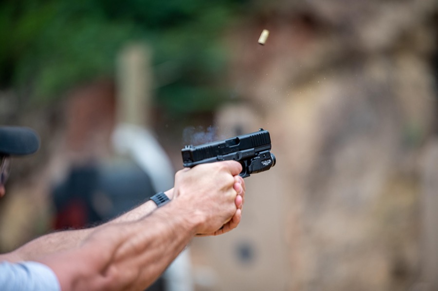 戶外靶場射擊特寫，雙手以拇指向前握法握持半自動手槍，彈殼正從拋殼口飛出