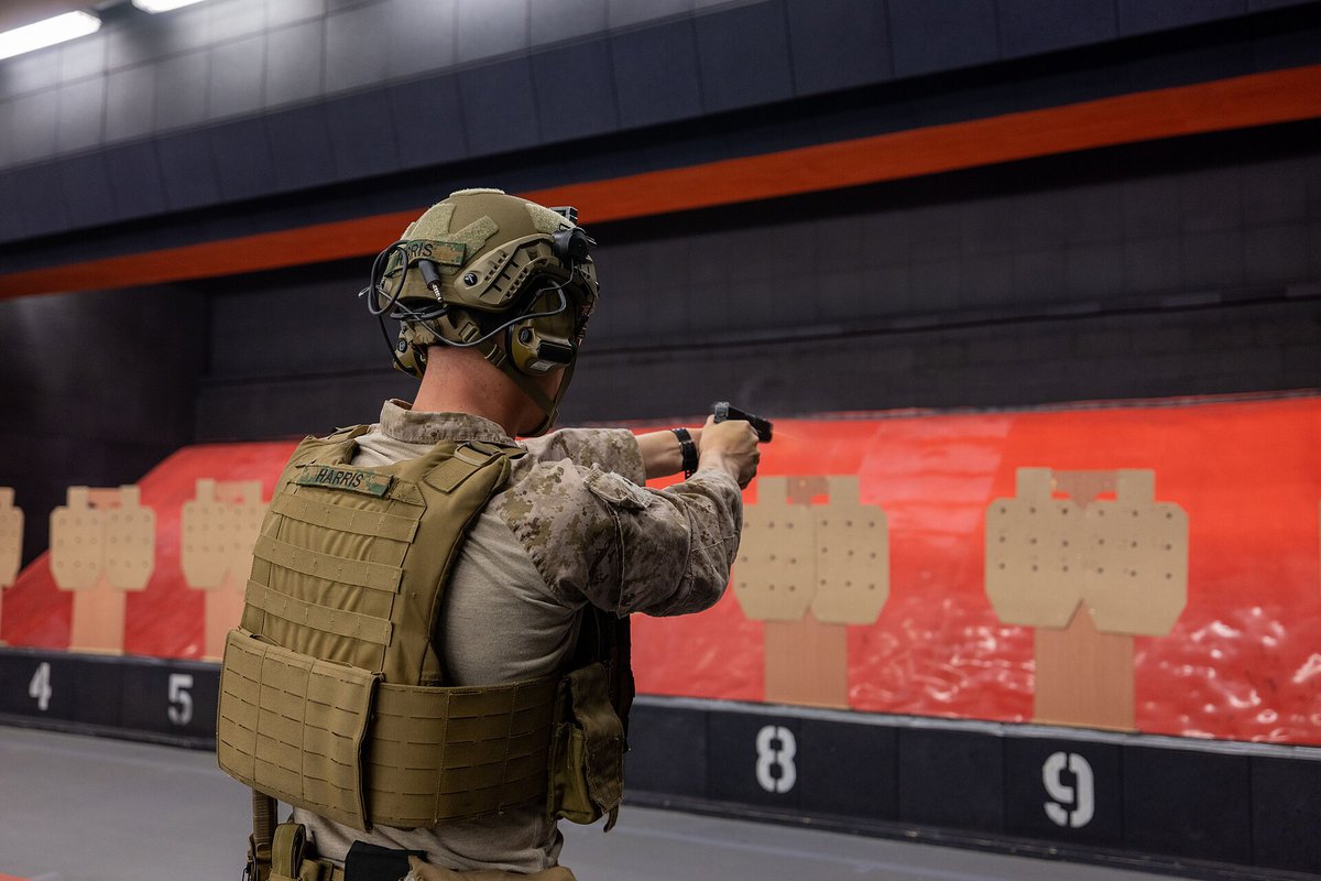 U.S. Marine in a plate carrier firing a pistol at cardboard targets a few meters away in an indoor range with a red backstop