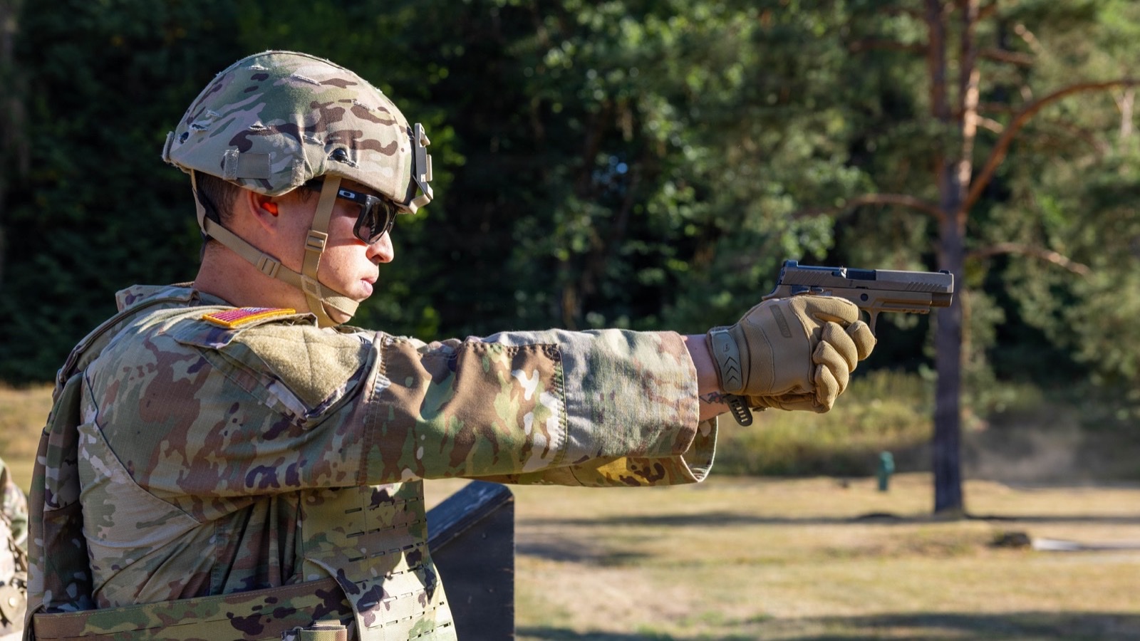 一名美國陸軍士兵戴著射擊眼鏡，在戶外靶場的手槍資格測驗中伸直手臂以 M17 手槍瞄準